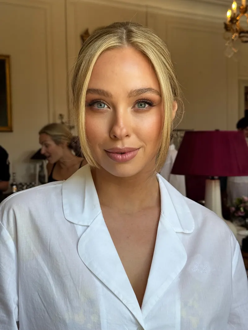 Blonde woman in white shirt with sleek tucked hair and glowing makeup, getting ready