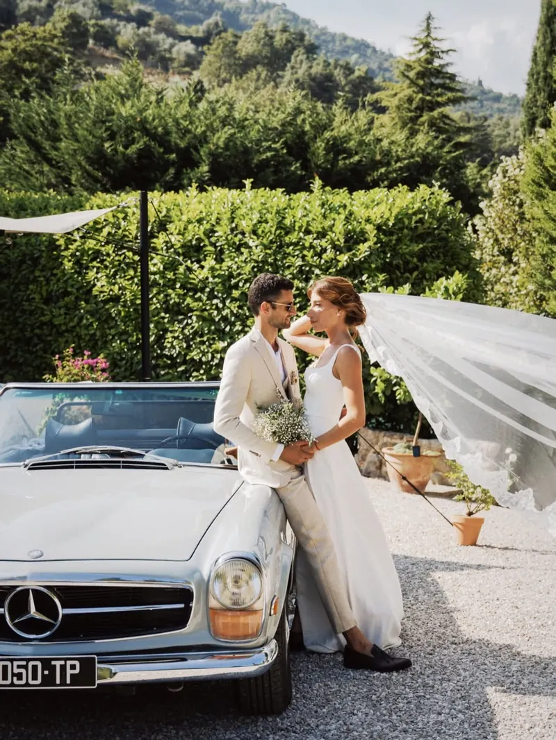Les mariés enlacés près d’un cabriolet vintage dans un jardin, voile au vent