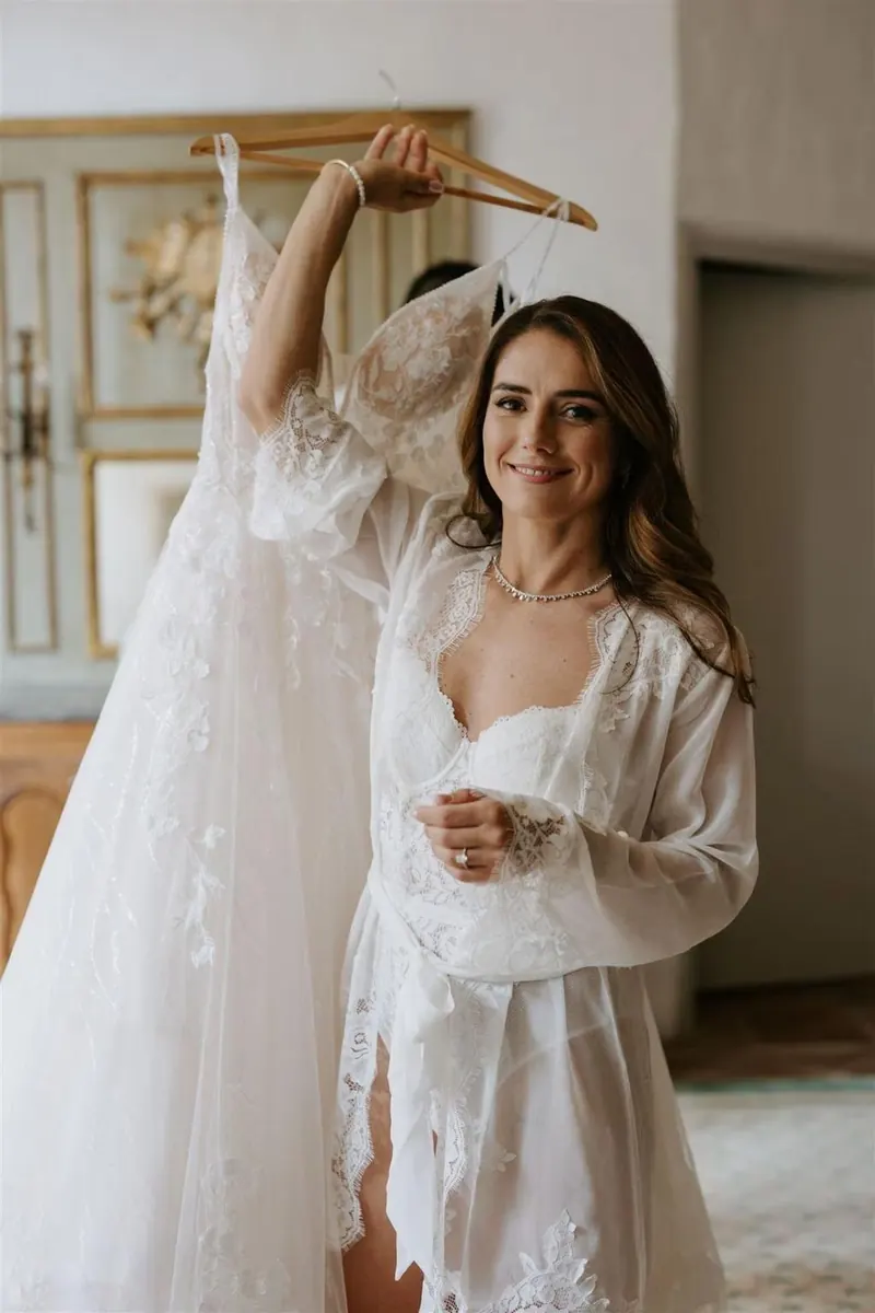 Bride in lace robe holding her wedding dress on a hanger while getting ready
