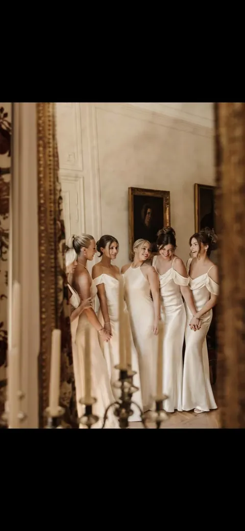 Five women in matching ivory satin gowns posing in an ornate salon, vertical crop
