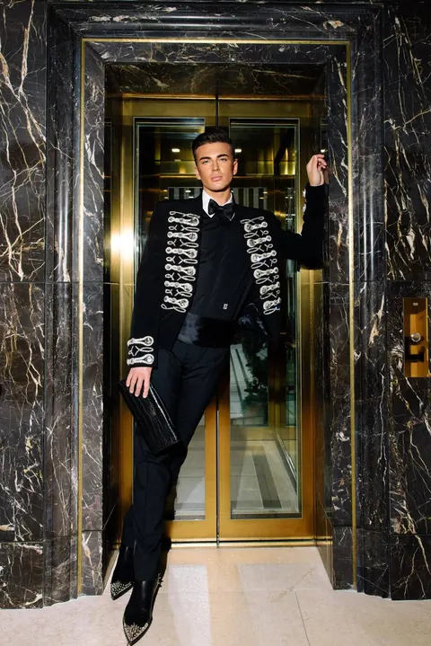 Male model in embroidered black jacket posing in a marble-framed lift doorway