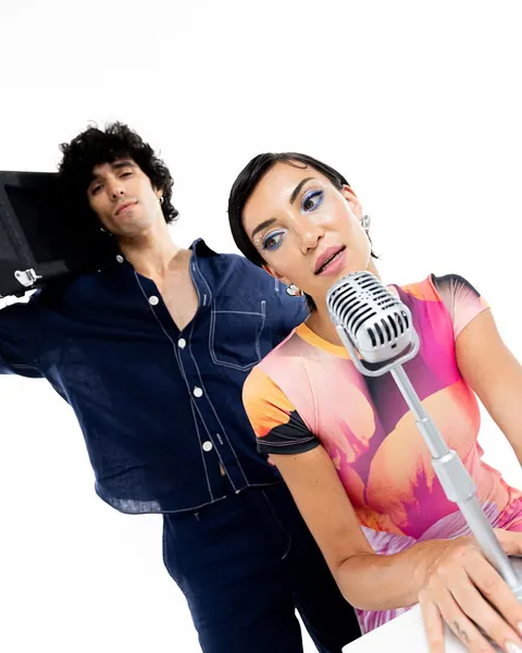 Man and woman with graphic blue eyeliner singing into a vintage microphone, studio