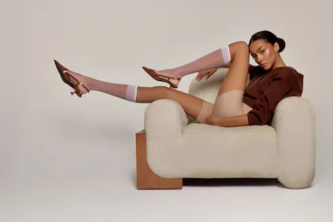 Model in brown sweater and sheer socks lounging on a boucle armchair, studio