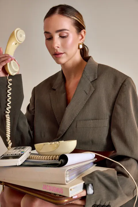 Model with sleek bun in an oversized blazer holding a retro phone and stack of books
