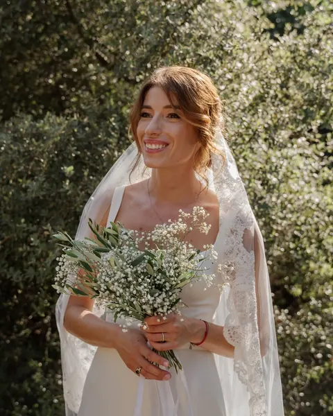 Smiling bride with lace veil and loose updo holding a gypsophila and olive bouquet