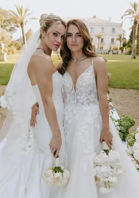 Two brides embracing on a villa lawn with palm trees, holding white bouquets