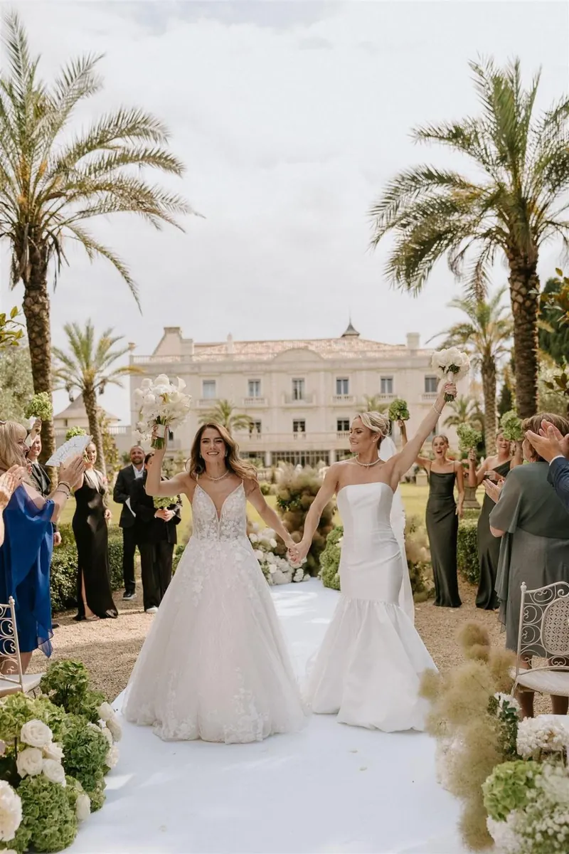 Two brides walking down a flower-lined aisle between palm trees as guests cheer