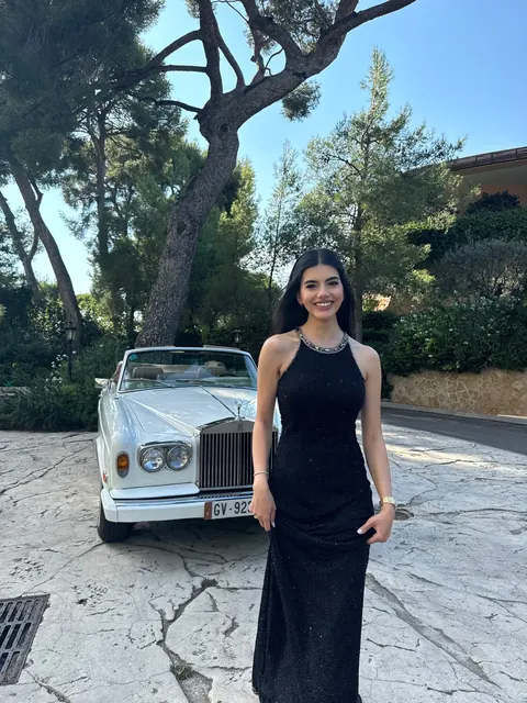 Woman in black sequined gown beside a vintage white car under pine trees
