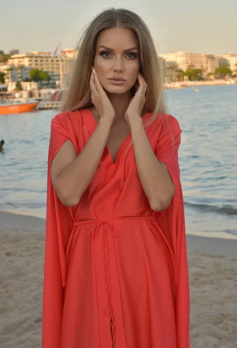 Woman in coral kaftan with hands framing her face on a beach, seafront behind