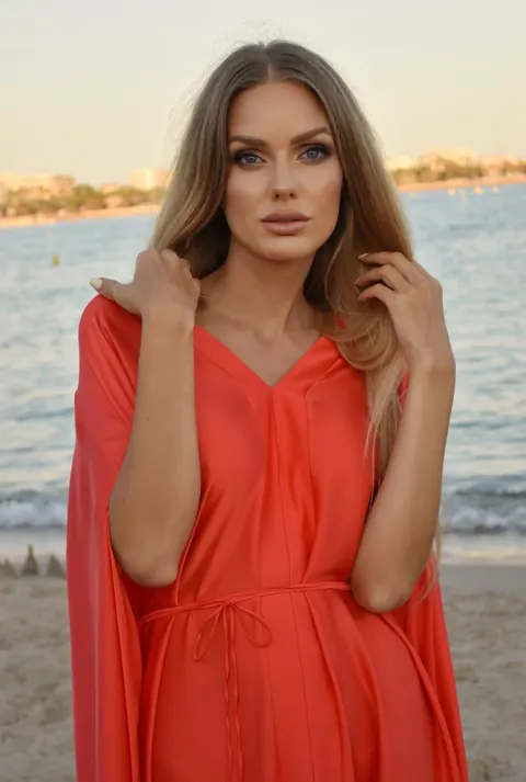 Woman in coral kaftan with long waves on a beach at sunset, sea behind