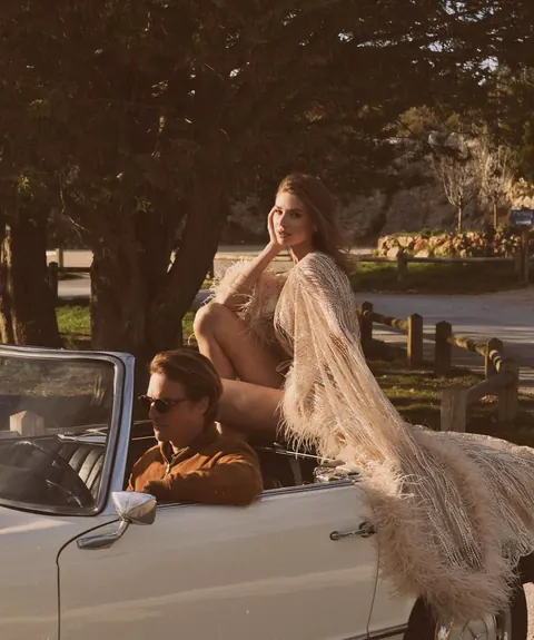 Woman in feathered fringe gown perched on a vintage convertible with driver
