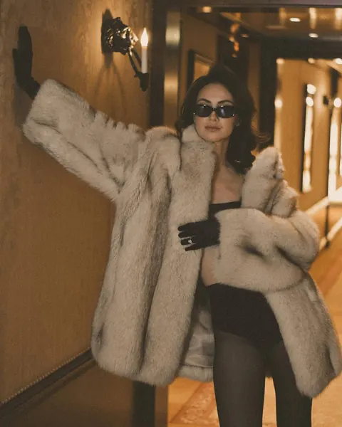 Woman in oversized fur coat and sunglasses posing in a dim hotel corridor