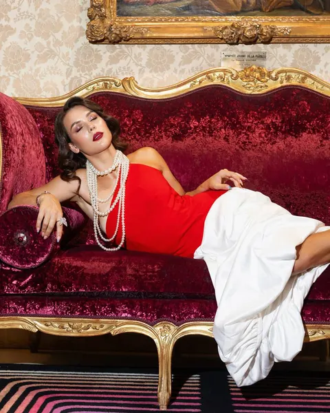 Woman in red dress and pearls lounging on a red velvet sofa, retro glam photoshoot
