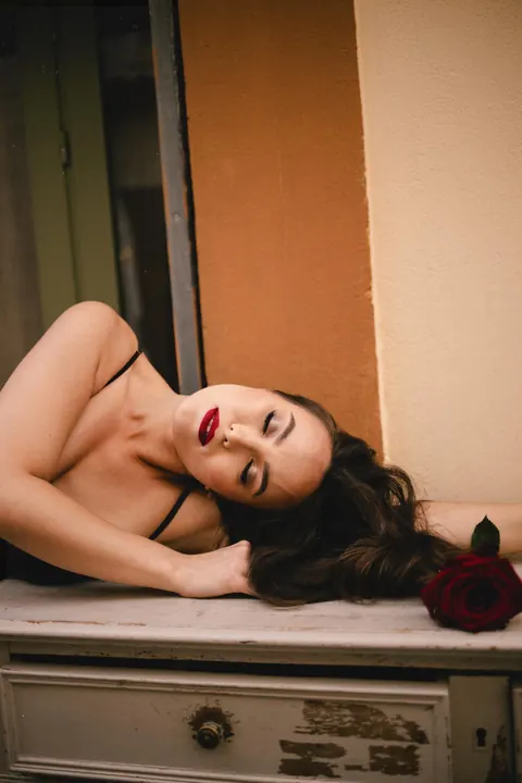 Woman with red lipstick lying on a vintage chest beside a red rose, moody photoshoot