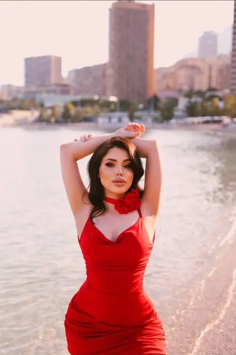 Woman in red satin dress with rose choker at the water's edge, high-rises behind