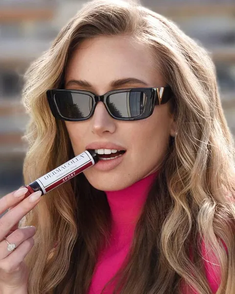 Woman in sunglasses with long beach waves applying a lip product, pink top