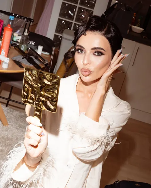 Woman in white feathered blazer blowing a kiss and holding a gold mirror, glam makeup