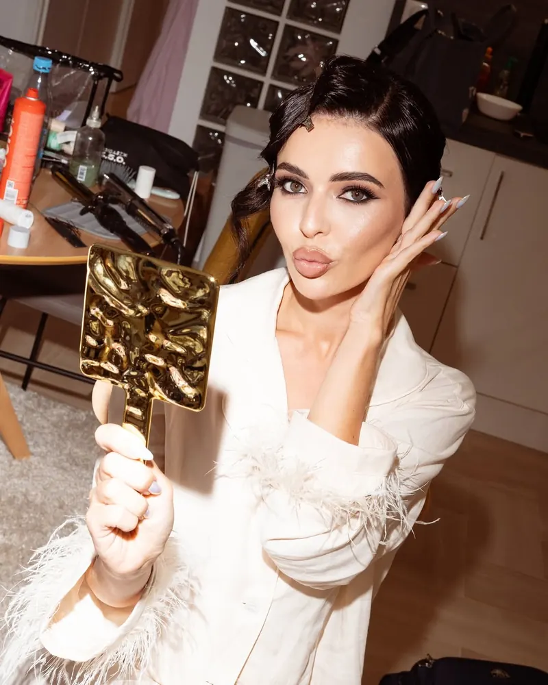 Woman in white feathered blazer blowing a kiss and holding a gold mirror, glam makeup